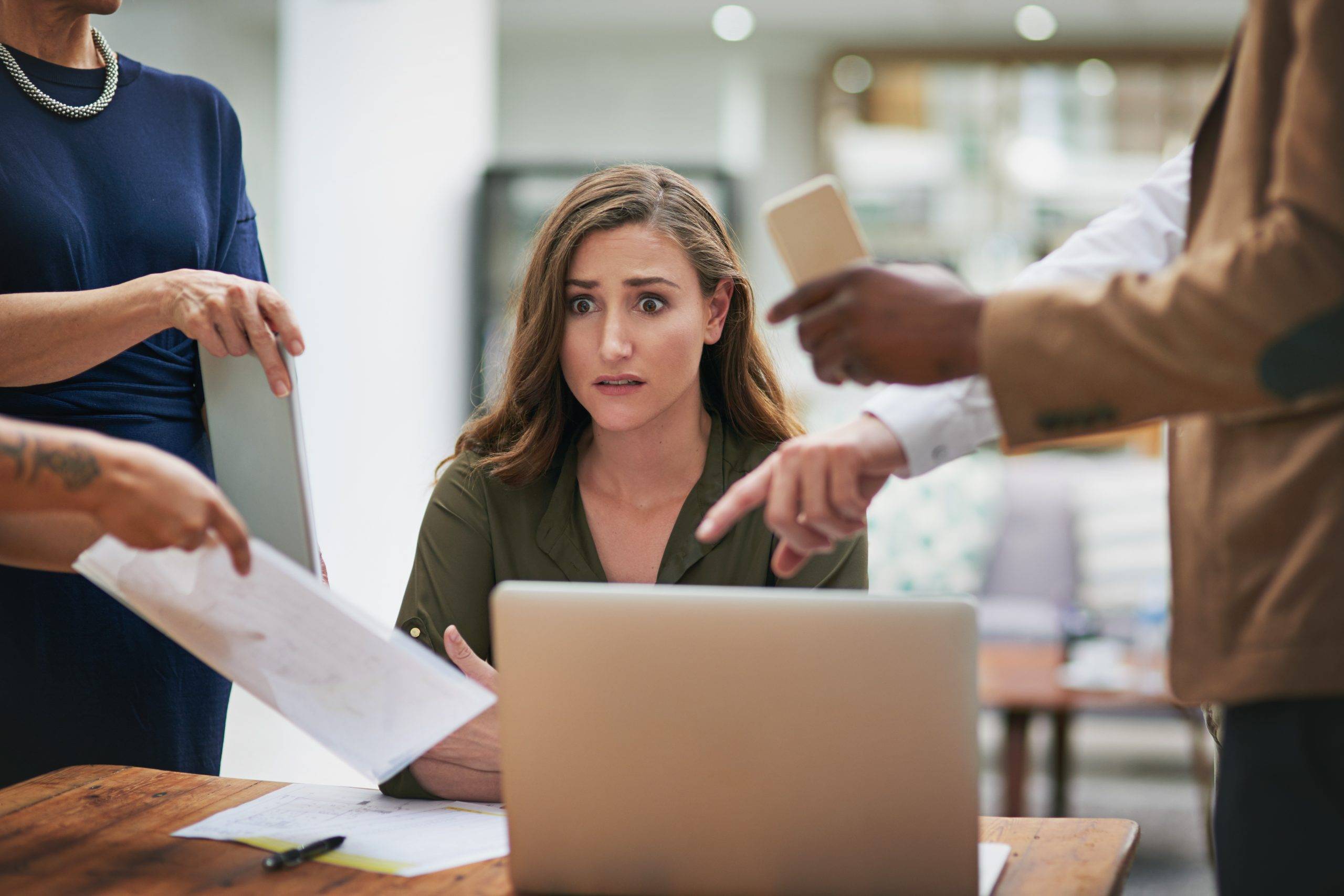 Employee burnout signs: A woman in a green shirt sits at her desk in front of a laptop, looking stressed as she juggles multiple tasks