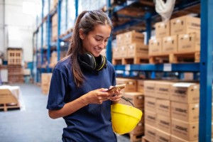 Young woman using her mobile phone in large warehouse. Female warehouse worker reading text message on her cell phone.