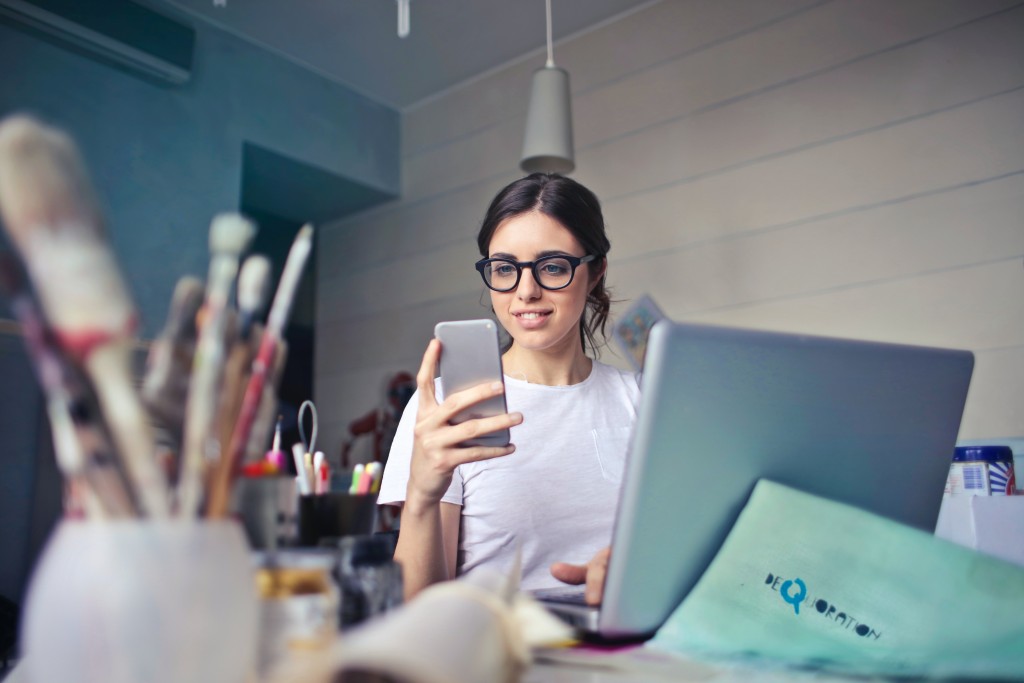 Employee communication - woman wearing glasses using smartphone and sitting in front of laptop computer