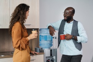 Happy young intercultural office workers chatting and having tea and coffee at break while standing by water dispenser with gallon