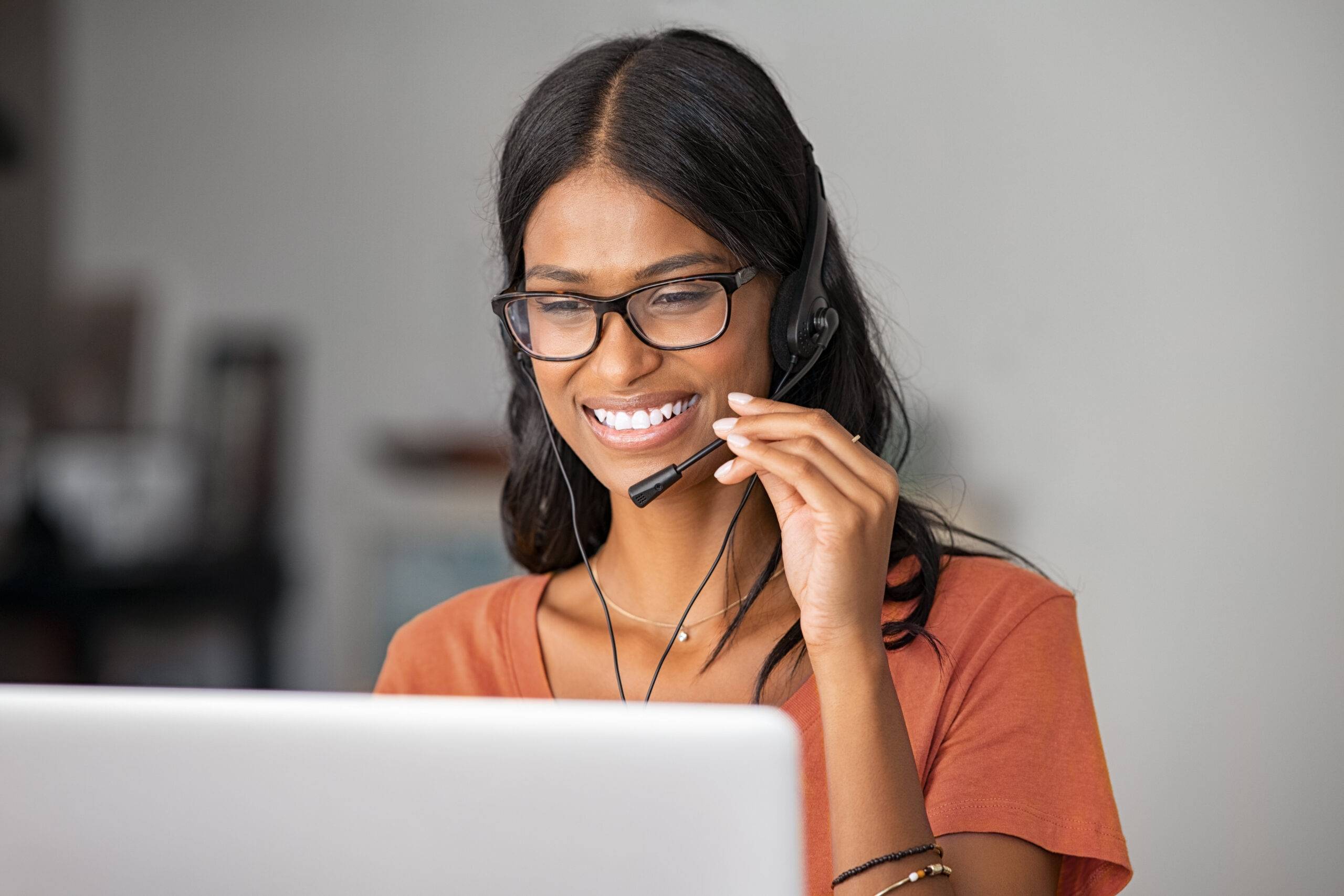 Net promoter score (NPS): A woman in an orange t-shirt wearing a headset, multitasking as she talks and types on her laptop, representing customer feedback and support.