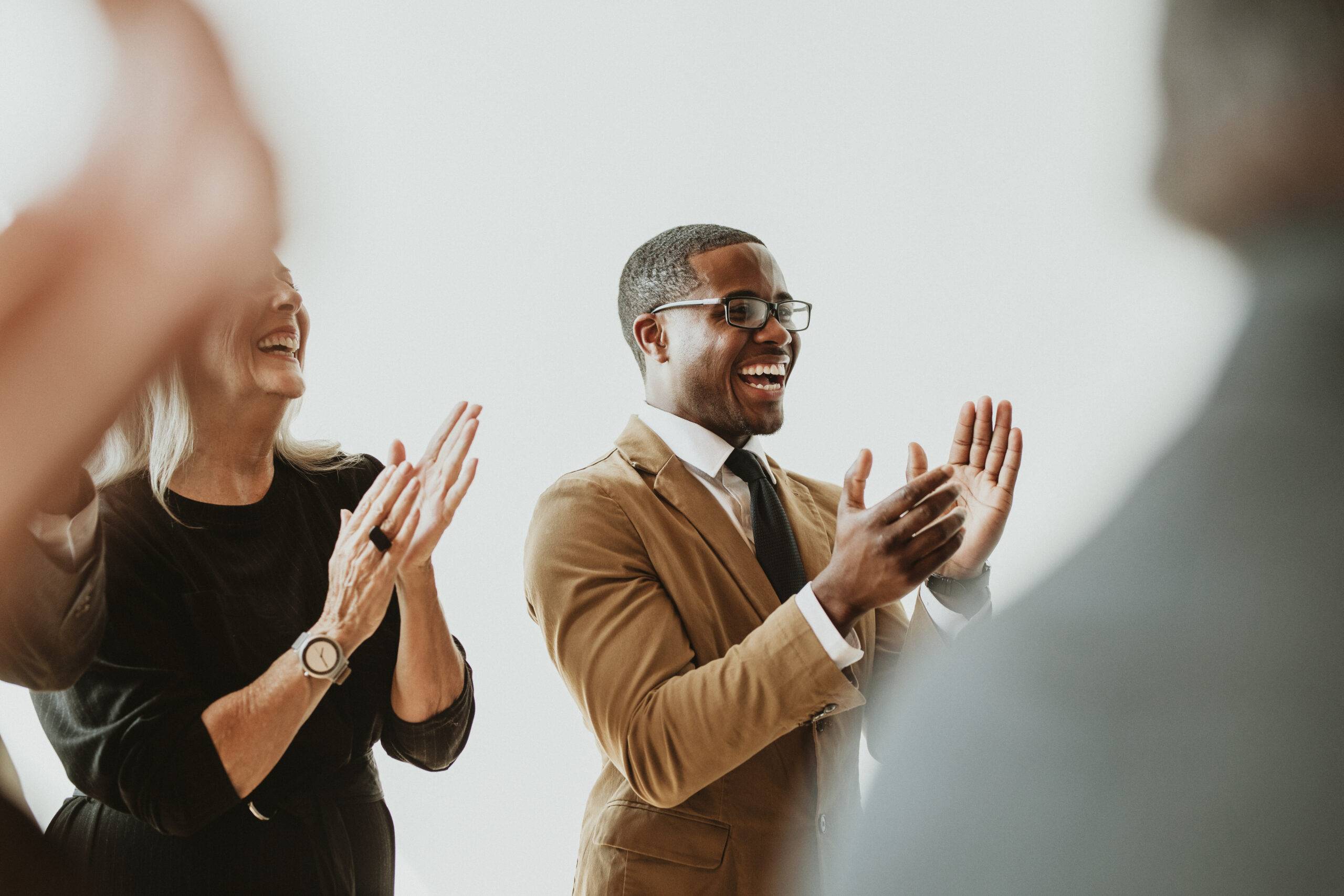 Employee recognition: A man in a brown suit and a woman in a black dress stand together, clapping in appreciation, with a blurred office setting in the background.