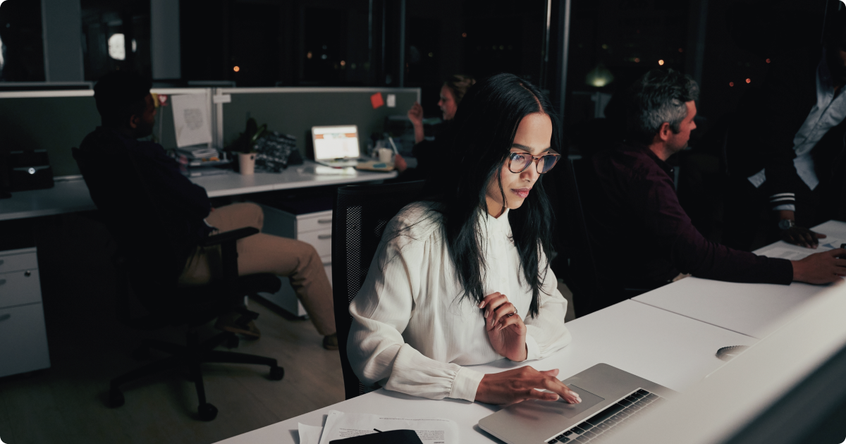 Organizational agility: A collaborative workspace where a woman in a white shirt wearing glasses is working on her laptop in a dim office setting.